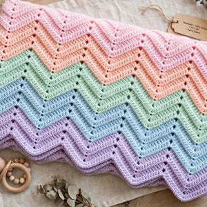 May include: A crochet blanket featuring a chevron pattern in pastel stripes of pink, peach, mint green, light blue, and lavender. A brown paper tag attached to the corner displays the text: Handmade - 100% Cotton - 30" x 36" - Soft & Gentle.