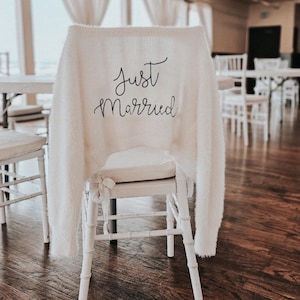 May include: A white chair with a cream-coloured, fluffy cover. The cover is embroidered with the words "Just Married" in black script. The chair is in a room with other white chairs and a wooden floor.