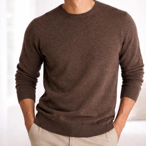 May include: A brown crew neck sweater with long sleeves. The sweater has a ribbed hem and cuffs. The person is wearing khaki pants. The sweater is made of a soft material.