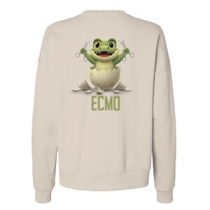 May include: Beige sweatshirt featuring a cartoon frog emerging from a cracked egg, holding scissors. The frog is green with a smiling expression. The text "ECMO" is printed in green below the egg.