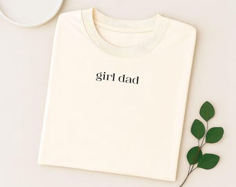 May include: Cream-colored t-shirt with the words "girl dad" printed in black. The shirt is folded neatly on a light beige surface, with a sprig of green leaves and a white plate in the background.