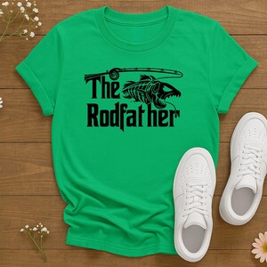 Puede incluir: Camiseta verde con el texto "The Rodfather" y un gr&aacute;fico negro de un esqueleto de pez con una ca&ntilde;a de pescar. Zapatillas blancas est&aacute;n junto a la camiseta sobre una superficie de madera. Tambi&eacute;n hay margaritas y peque&ntilde;as flores blancas.