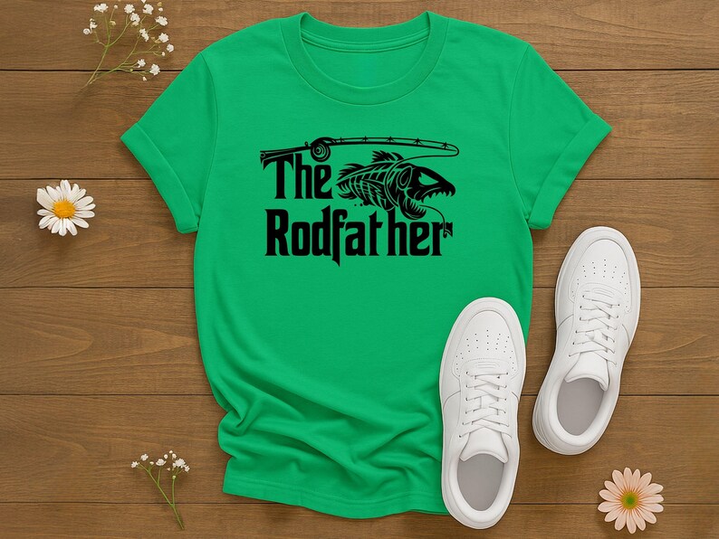 Puede incluir: Camiseta verde con el texto "The Rodfather" y un gr&aacute;fico negro de un esqueleto de pez con una ca&ntilde;a de pescar. Zapatillas blancas est&aacute;n junto a la camiseta sobre una superficie de madera. Tambi&eacute;n hay margaritas y peque&ntilde;as flores blancas.