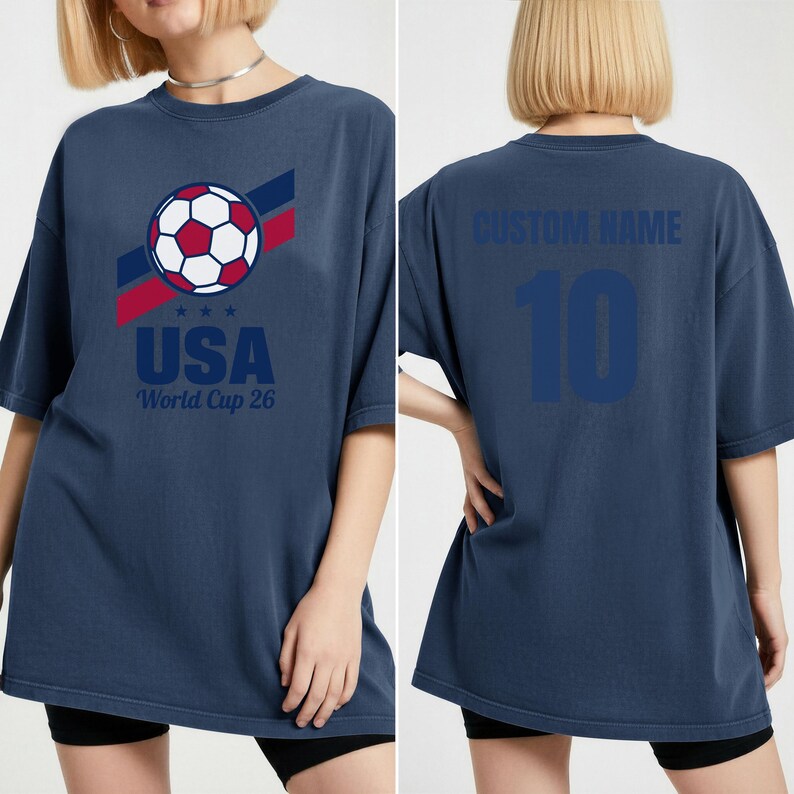 Puede incluir: Camiseta azul marino con un bal&oacute;n de f&uacute;tbol, rayas rojas y azules, y el texto "USA World Cup 26". La parte trasera de la camiseta muestra "CUSTOM NAME" sobre el n&uacute;mero "10" en azul.