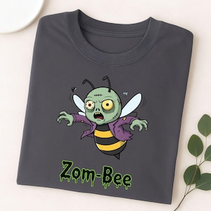 May include: Dark grey t-shirt with a cartoon zombie bee design. The bee has green skin, a purple jacket, and yellow and black stripes. The text "Zom-Bee" is in green, dripping letters. The design is centered on the shirt.