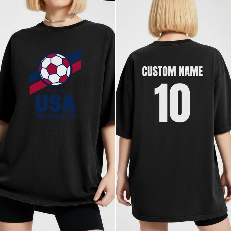 Puede incluir: Camiseta negra con un dise&ntilde;o gr&aacute;fico de bal&oacute;n de f&uacute;tbol, rayas rojas y azules y el texto "USA World Cup 26". La parte trasera muestra "CUSTOM NAME" sobre el n&uacute;mero "10" en blanco. Una prenda deportiva y casual.