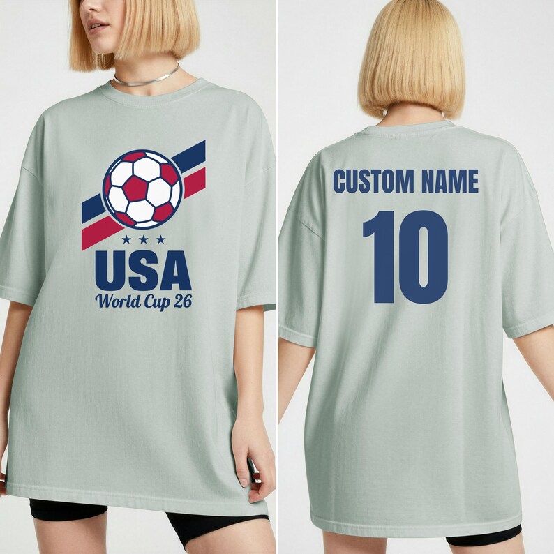 Puede incluir: Camiseta verde claro con un gr&aacute;fico de bal&oacute;n de f&uacute;tbol y el texto "USA World Cup 26". La parte trasera de la camiseta tiene el texto "CUSTOM NAME" encima del n&uacute;mero "10" en azul marino. La camiseta es de corte holgado.
