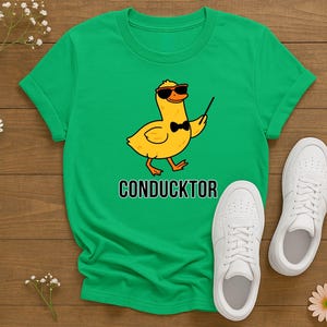 T-shirt featuring a cartoon duck wearing sunglasses and a bow tie, holding a conductor’s baton, with the word “CONDUCKTOR” printed below as a music pun.
