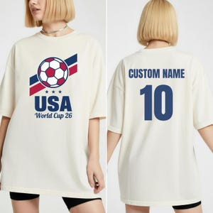 Puede incluir: Camiseta color crema con un gr&aacute;fico de bal&oacute;n de f&uacute;tbol, texto USA y "World Cup 26". La parte trasera de la camiseta tiene "CUSTOM NAME" y el n&uacute;mero 10 en azul. La camiseta tiene un corte holgado y oversize.