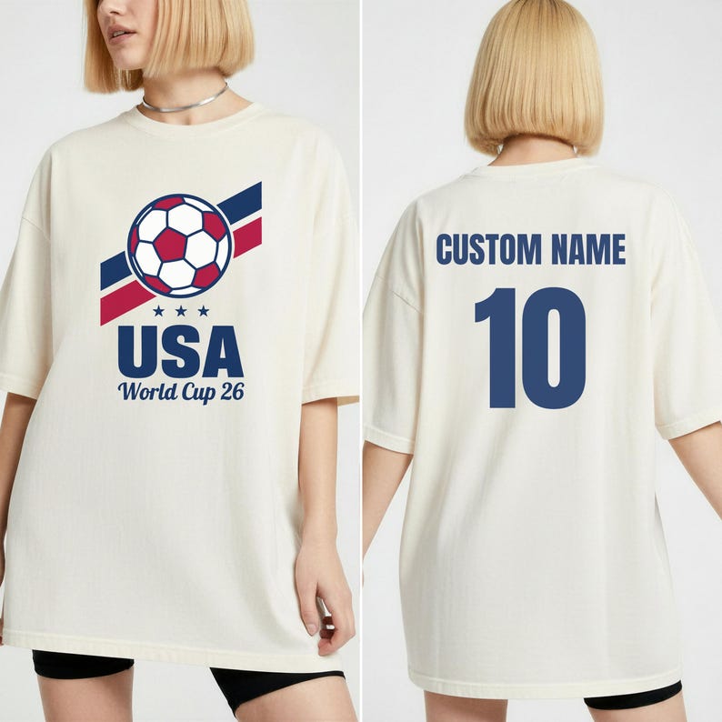 Puede incluir: Camiseta color crema con un gr&aacute;fico de bal&oacute;n de f&uacute;tbol, texto USA y "World Cup 26". La parte trasera de la camiseta tiene "CUSTOM NAME" y el n&uacute;mero 10 en azul. La camiseta tiene un corte holgado y oversize.