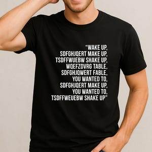 May include: Black t-shirt with white text. The text reads "WAKE UP, SDFGHJQERT MAKE UP, TSDFFWUEBW SHAKE UP, WQEFZDVRG TABLE, SDFGHJQWERT FABLE, YOU WANTED TO, SDFGHJQERT MAKE UP, YOU WANTED TO, TSDFFWEUEBW SHAKE UP."