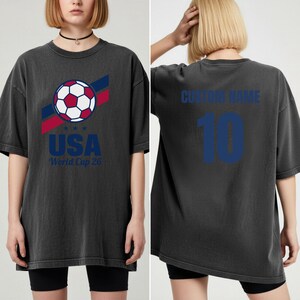 Puede incluir: Camiseta gris oscuro con un dise&ntilde;o de bal&oacute;n de f&uacute;tbol, rayas rojas y azules, y el texto "USA" y "World Cup 26". La parte trasera muestra "CUSTOM NAME" y el n&uacute;mero "10" en azul.