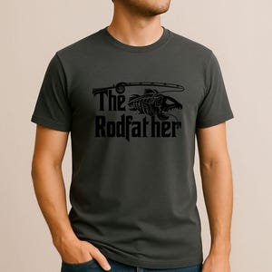 Puede incluir: Camiseta gris oscuro con un gr&aacute;fico negro de un esqueleto de pez y una ca&ntilde;a de pescar. El texto "The Rodfather" est&aacute; debajo en una fuente estilizada. La camiseta es de material suave.