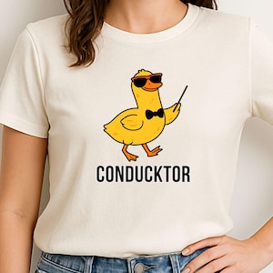 Może przedstawiać: Biały t-shirt z żółtą kaczką w okularach przeciwsłonecznych i muszce, trzymającą dyrygentkę. Pod kaczką znajduje się napis "CONDUCKTOR".