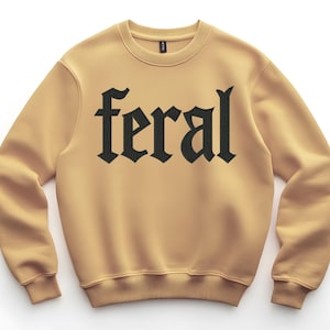 Op de afbeelding: Een lichtbruine sweatshirt met het woord "feral" in grote, zwarte gotische letters op de borst. De sweatshirt heeft een ronde hals en lange mouwen met geribde manchetten en zoom.