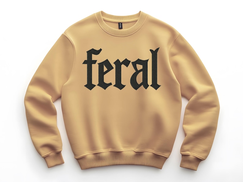 Op de afbeelding: Een lichtbruine sweatshirt met het woord "feral" in grote, zwarte gotische letters op de borst. De sweatshirt heeft een ronde hals en lange mouwen met geribde manchetten en zoom.