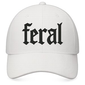 Op de afbeelding: Witte baseballcap met het woord "feral" geborduurd in zwarte gotische letters. De cap heeft een gebogen klep en een knoop aan de bovenkant. Een casual accessoire.