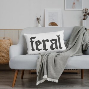 Op de afbeelding: Een wit rechthoekig kussen met het woord "feral" in zwarte gotische letters. Het kussen ligt op een lichtgrijze bank met een grijze deken eroverheen. Een gevlochten mand en ingelijste kunstwerken zijn op de achtergrond.