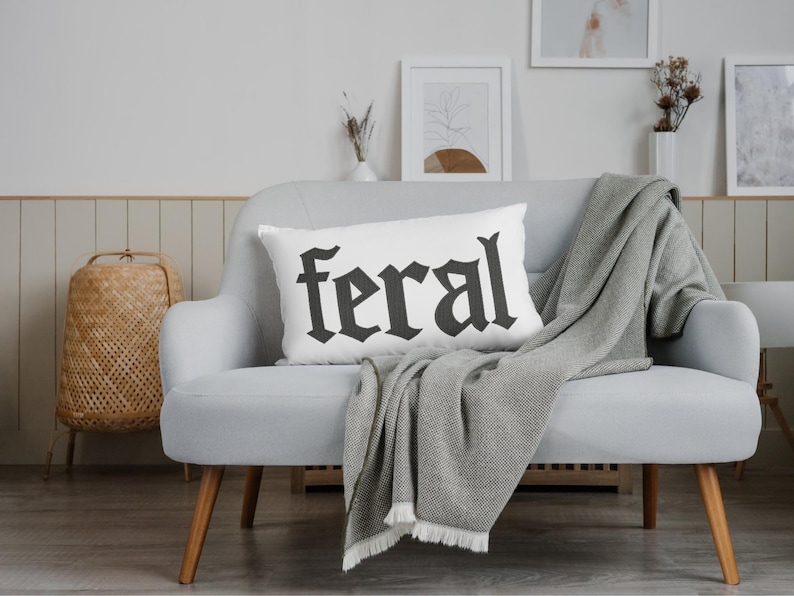 Op de afbeelding: Een wit rechthoekig kussen met het woord "feral" in zwarte gotische letters. Het kussen ligt op een lichtgrijze bank met een grijze deken eroverheen. Een gevlochten mand en ingelijste kunstwerken zijn op de achtergrond.
