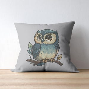 May include: A decorative gray pillow featuring a watercolor illustration of a cartoon owl perched on a branch. The owl has large eyes, blue and tan feathers, and is surrounded by green leaves. The pillow is on a wooden surface.