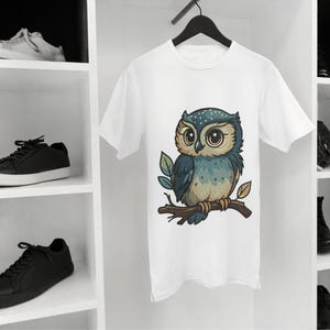 May include: White t-shirt featuring a cartoon owl perched on a branch. The owl has large eyes, blue and tan feathers, and is set against a white background. The shirt is hanging on a black hanger.