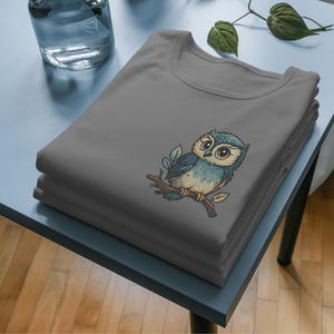 May include: A stack of gray t-shirts featuring an embroidered owl design. The owl is perched on a branch with leaves, and has blue, brown, and white coloring. The shirts are neatly folded on a light blue table.