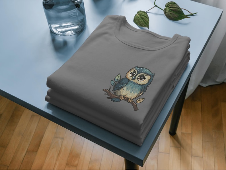 May include: A stack of gray t-shirts featuring an embroidered owl design. The owl is perched on a branch with leaves, and has blue, brown, and white coloring. The shirts are neatly folded on a light blue table.