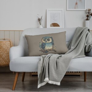 May include: A rectangular decorative pillow with a cartoon owl illustration. The owl is perched on a branch with leaves, featuring blue, beige, and brown colors. The pillow is on a white couch with a gray blanket.