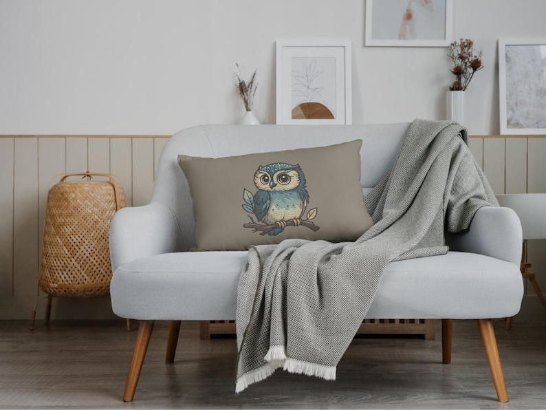 May include: A rectangular decorative pillow with a cartoon owl illustration. The owl is perched on a branch with leaves, featuring blue, beige, and brown colors. The pillow is on a white couch with a gray blanket.