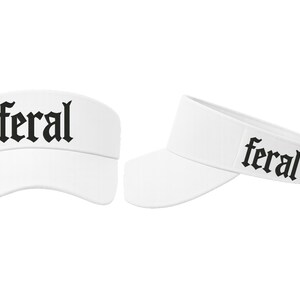 Op de afbeelding: Twee witte vizieren met het woord "feral" in zwarte gotische letters. De vizieren worden vanuit verschillende hoeken getoond, &eacute;&eacute;n recht van voren en de andere in een lichte hoek, waarbij het ontwerp wordt benadrukt.