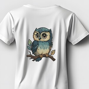 May include: White t-shirt featuring a cartoon owl illustration. The owl is perched on a brown branch with green and brown leaves. The owl has large eyes, blue and tan feathers, and is centered on the back of the shirt.
