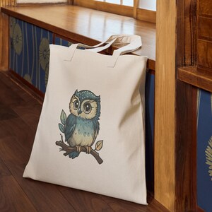 May include: A cream-colored tote bag featuring a detailed illustration of a blue and tan owl perched on a branch with leaves. The owl has large, expressive eyes. The bag is made of a sturdy fabric and has two handles.