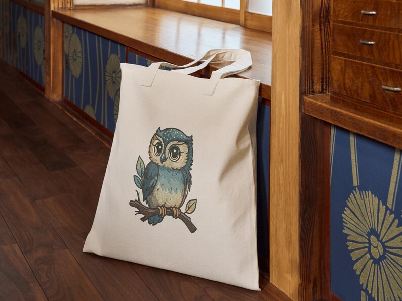 May include: A cream-colored tote bag featuring a detailed illustration of a blue and tan owl perched on a branch with leaves. The owl has large, expressive eyes. The bag is made of a sturdy fabric and has two handles.