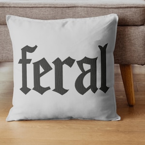 Op de afbeelding: Een wit kussen met het woord "feral" geborduurd in donkergrijze gotische letters. Het kussen ligt op een houten vloer voor een grijze bank.