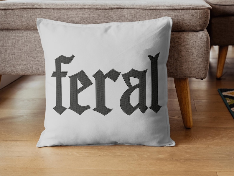 Op de afbeelding: Een wit kussen met het woord "feral" geborduurd in donkergrijze gotische letters. Het kussen ligt op een houten vloer voor een grijze bank.
