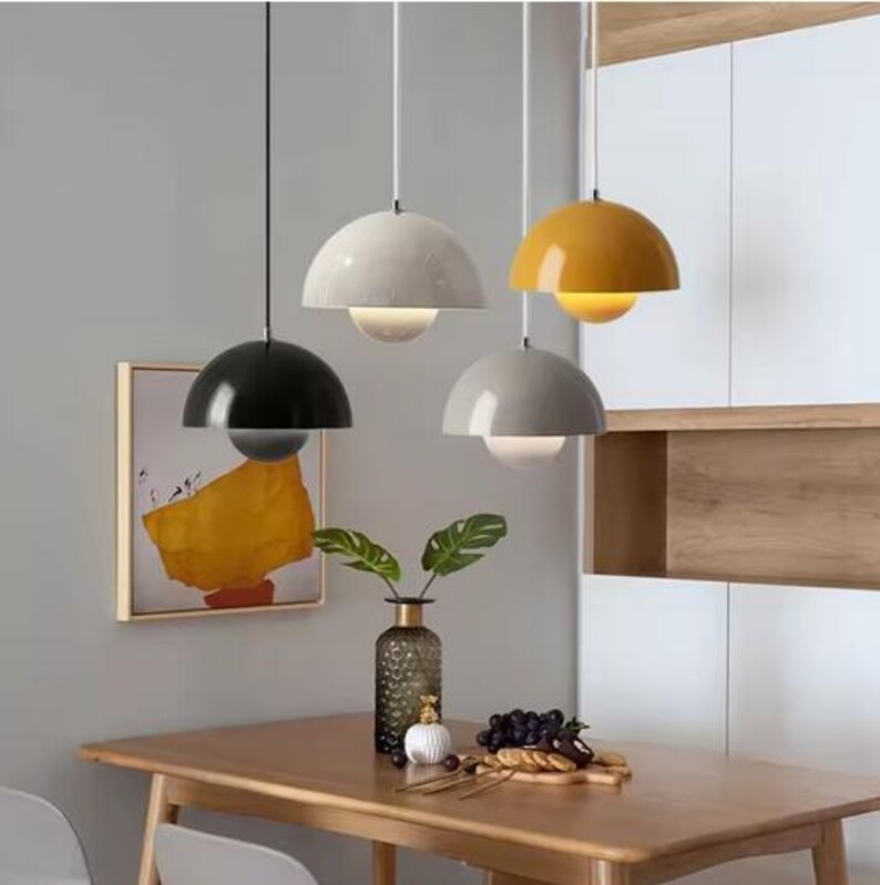 May include: Four modern pendant lights with a semi-spherical shade in black, white, gray, and yellow. Each light has a spherical glass bulb. The lights hang above a wooden table with a vase of greenery and a framed abstract painting.