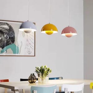 May include: Three pendant lights with a modern design. Each light has a rounded, colored shade: light blue, yellow, and pink. The lights hang above a white table with a vase of flowers and a framed artwork on the wall.