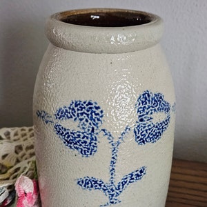 May include: A vintage stoneware crock with a cream-colored base and a dark brown interior. The crock features a blue floral design. The jar has a textured surface and a rounded top. A classic piece for kitchen or home decor.
