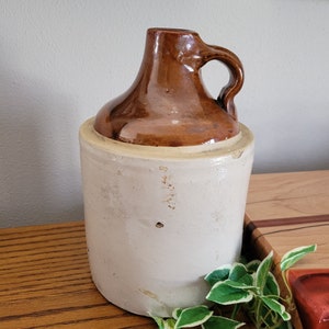 May include: A vintage stoneware jug with a brown top and handle, and a cream-colored body. The jug has a cylindrical shape and a rounded top. It is sitting on a wooden surface.