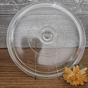 May include: A circular, clear glass lid featuring a raised, centered knob handle. The lid has a stepped, concentric ring design on its surface, with a smooth, rounded outer rim. The glass is transparent, allowing a clear view through the object.
