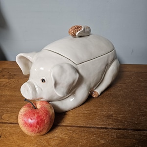 May include: A white ceramic pig-shaped container with a corn cob lid. The pig has detailed features, including ears, snout, and eyes. A red and yellow apple rests beside the pig on a wooden surface.