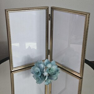 May include: A double hinged picture frame with a metallic gold finish and a beaded edge detail. The frame features two rectangular vertical openings with white inserts, connected by a central metal hinge, and is positioned on a reflective surface.