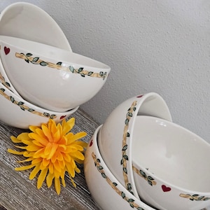 May include: A stack of white ceramic bowls with a decorative border of a brown branch and green leaves. Each bowl has a red heart design. A bright yellow flower is in the foreground.