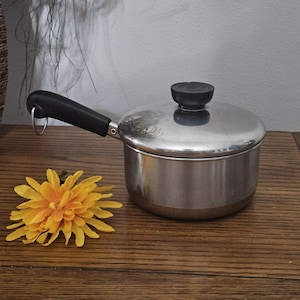 May include: A small, stainless steel saucepan with a black handle and lid. The handle has a metal ring for hanging. A yellow and orange flower is on the wooden surface next to the pan.