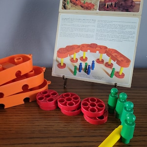 May include: An orange and red vintage toy set with a colorful design. The set includes orange boat-shaped pieces, red wheels, green figures, and a yellow marker. The box shows the toy in use. The text on the box reads "THE AMB TOY IS A COLORFUL, SAFE FUN TOY WHICH GROWS WITH A CH".