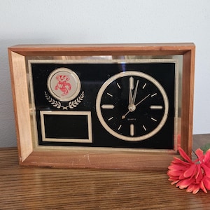 May include: A rectangular wooden clock with a black face and gold accents. The clock features a round clock face, a logo, and a blank rectangular space. The clock is a quartz movement.
