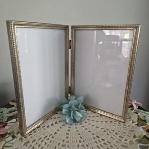May include: A double hinged picture frame with a metallic gold finish and a beaded edge detail. The frame consists of two rectangular panels connected by metal hinges, each containing a white insert, and is accented by a small blue flower.