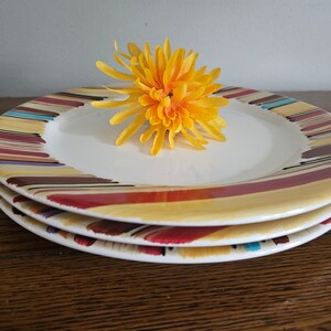 May include: A stack of three round ceramic plates with a white center and a wide rim decorated with multi-colored brushstroke patterns in shades of red, yellow, brown, and blue. A bright yellow artificial flower rests on the top plate.