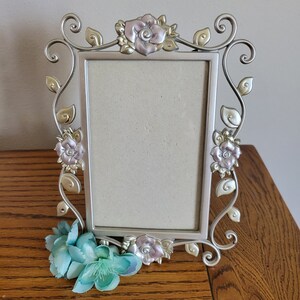 May include: A rectangular silver-toned metal picture frame featuring decorative scrollwork and sculpted floral accents in soft pink and cream hues. The frame is adorned with metal leaves and vines, and a cluster of artificial turquoise flowers sits at the base.