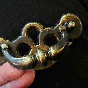 May include: Antique brass drawer pull with a decorative design. The pull features a curved handle and two circular backplates. The brass has a slightly aged appearance, adding to its vintage charm. Ideal for furniture restoration or adding a classic touch.